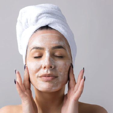 Mulher com toalha no cabelo a aplicar um produto de limpeza no rosto, num momento de rotina de skincare e cuidado facial.